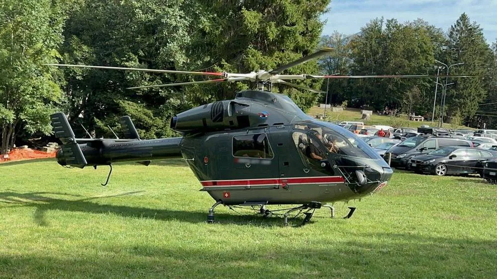 A dark grey helicopter with a red stripe on its side is parked on a grassy field, with trees and cars in the background.