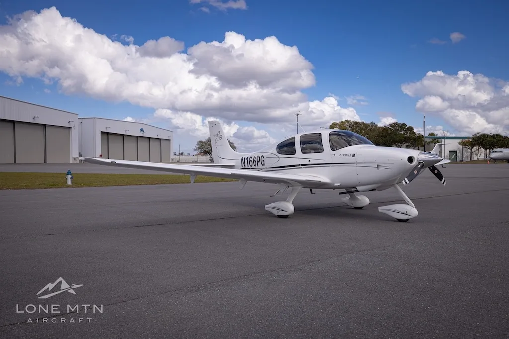 A white Cirrus SR20 aircraft (N166PG) is parked on an airport tarmac with hangars under a partly cloudy sky.