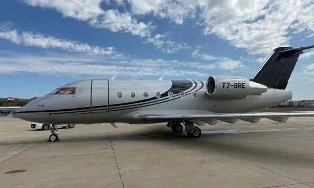 A silver Bombardier Challenger business jet with black and white stripes, registration T7-BRE, parked on an airfield under a partly cloudy sky.