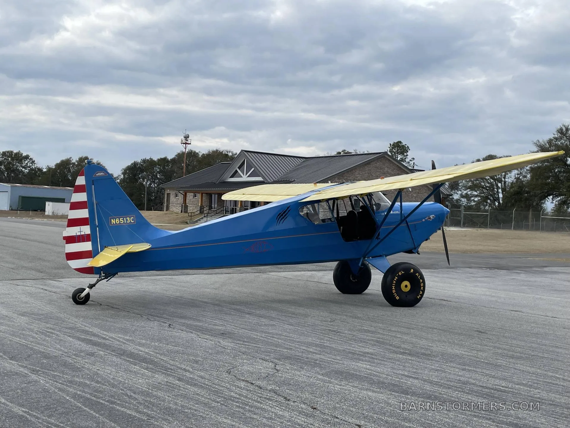 A blue and yellow high-wing aircraft with large tundra tires, registration N6513C, parked on an airfield with a building in the background.