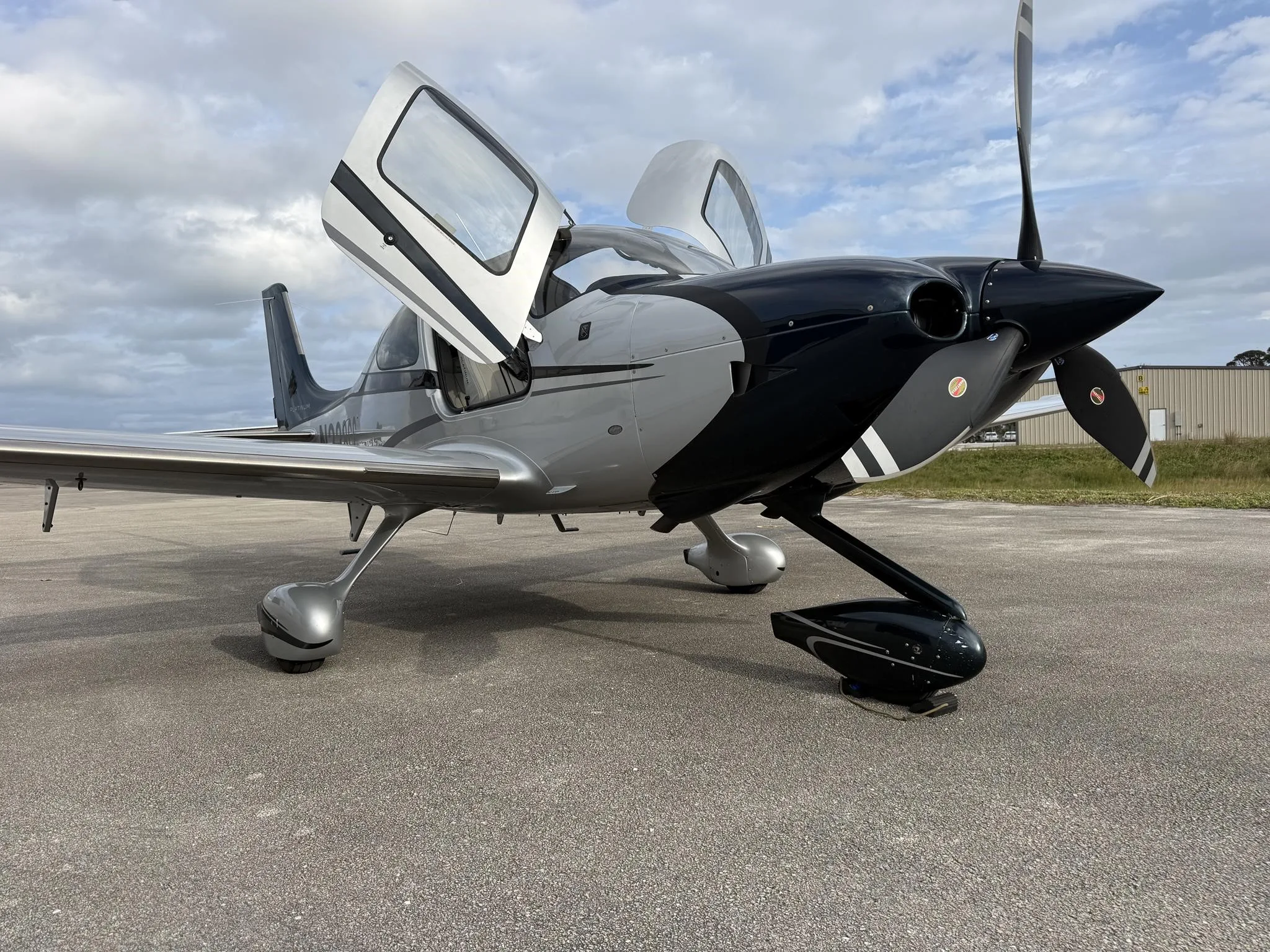 A silver and black Cirrus SR22 aircraft with its doors open, parked on an asphalt surface under a cloudy sky.