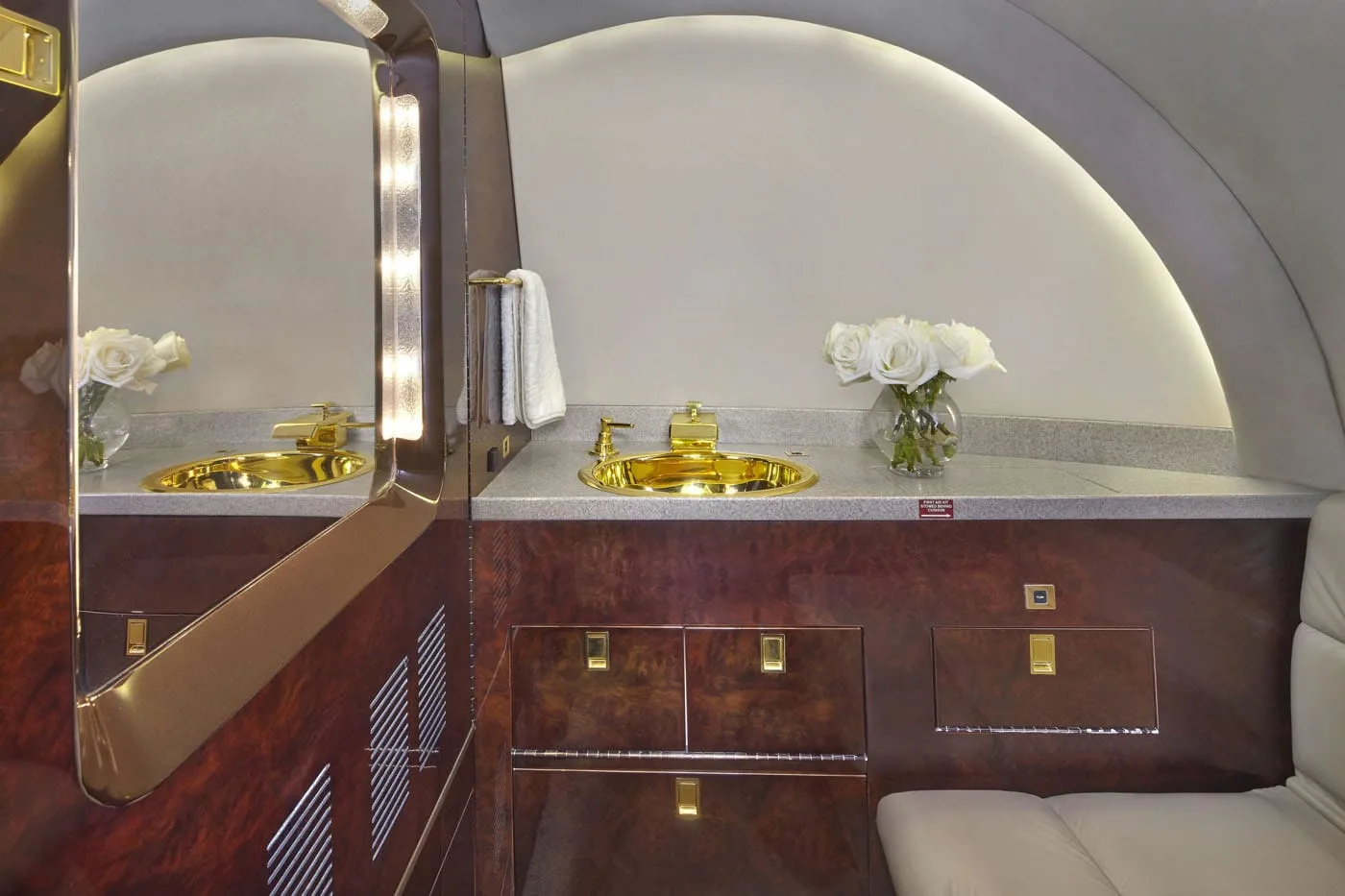 A luxurious aircraft lavatory with a gold sink, dark wood cabinetry, and a large mirror with vertical lighting.