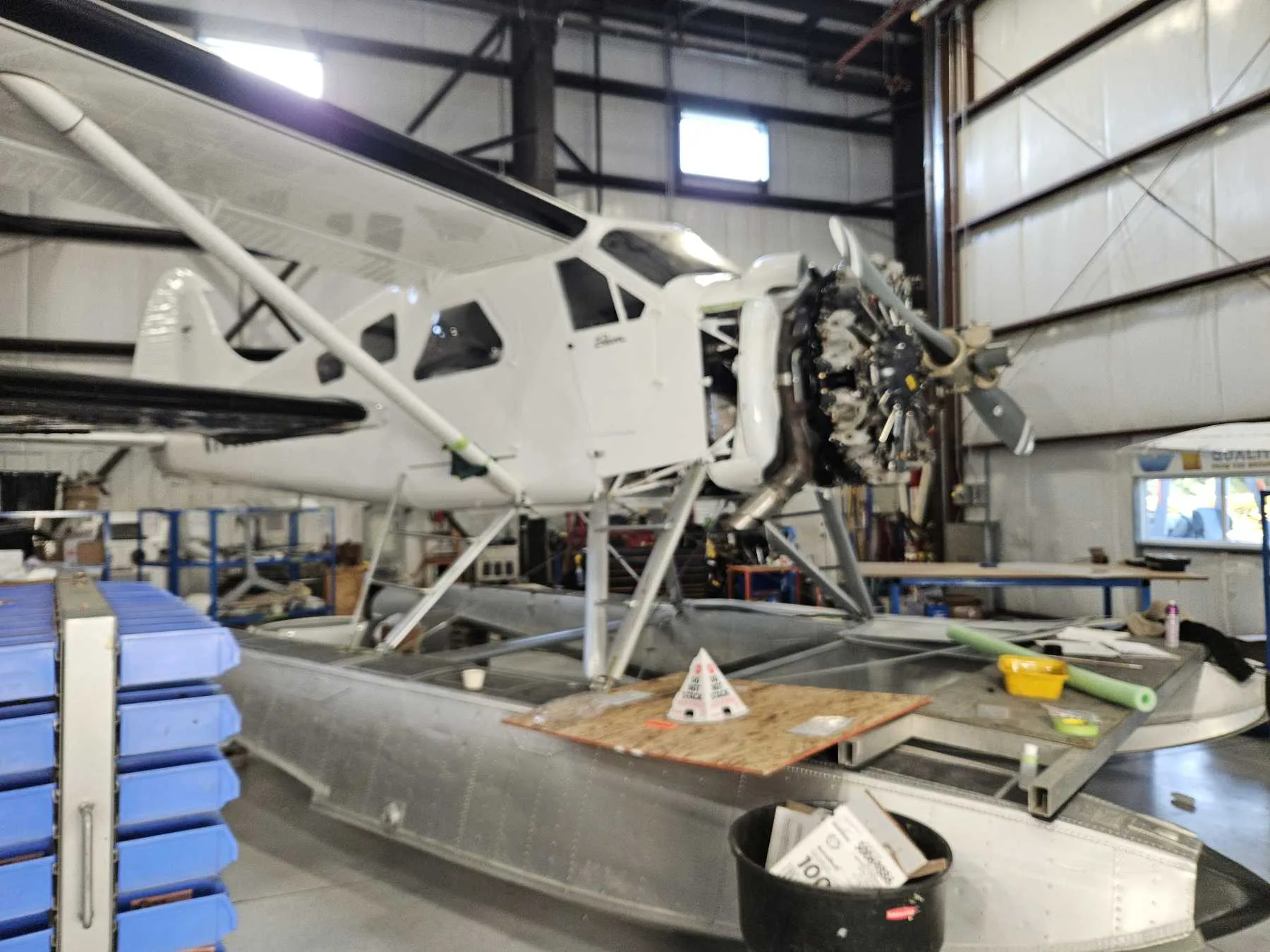 A white floatplane with its engine exposed, undergoing maintenance in a hangar.