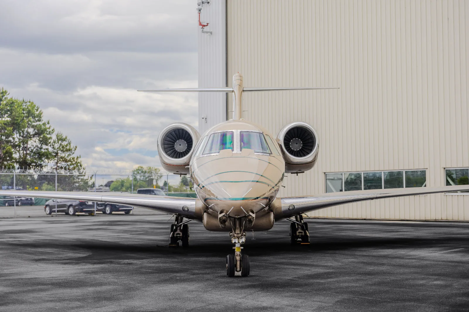 A front view of a beige business jet with green stripes parked on an asphalt tarmac in front of a hangar.
