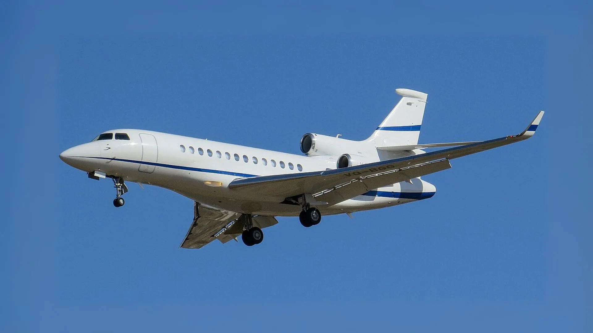 A white and blue Dassault Falcon business jet is shown in flight against a clear blue sky.