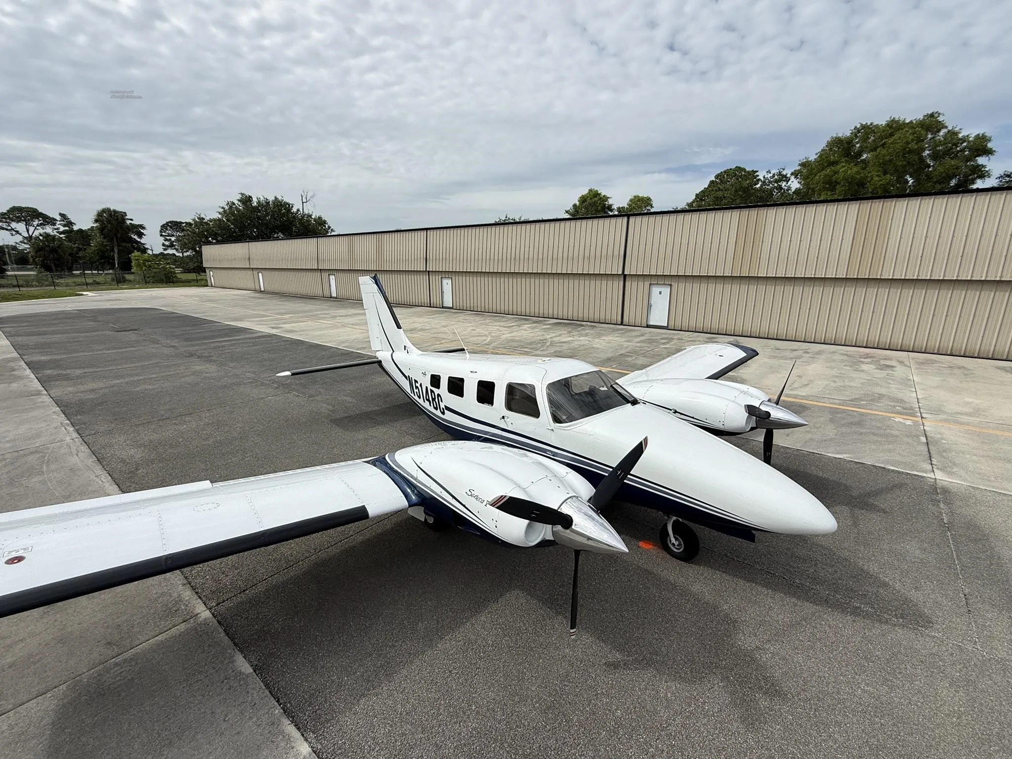A white and blue Piper Seneca V aircraft, registration N514BC, parked on an airfield tarmac with hangars in the background.