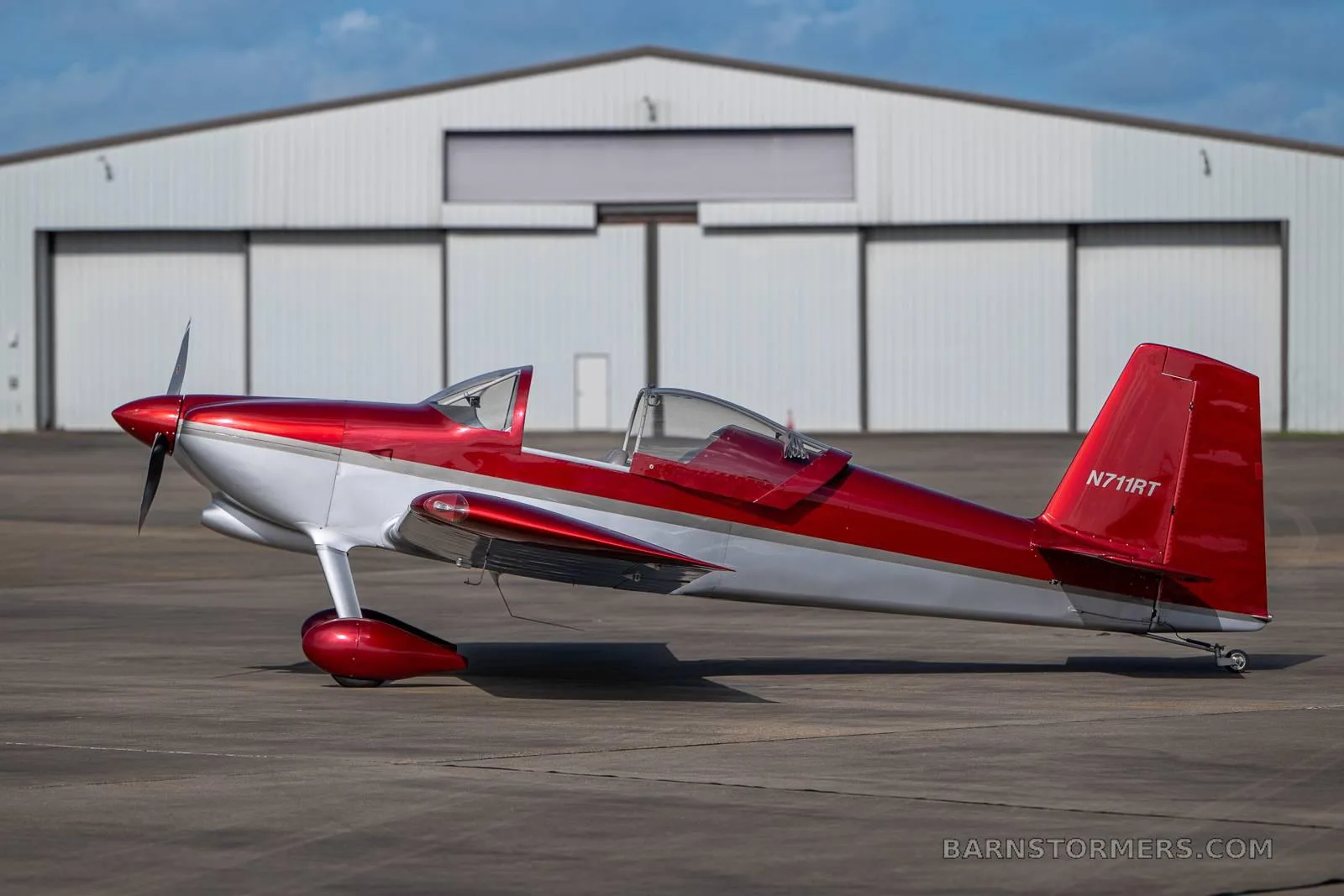 A red and silver single-engine experimental aircraft, N711RT, is parked in profile on a tarmac in front of a white hangar.