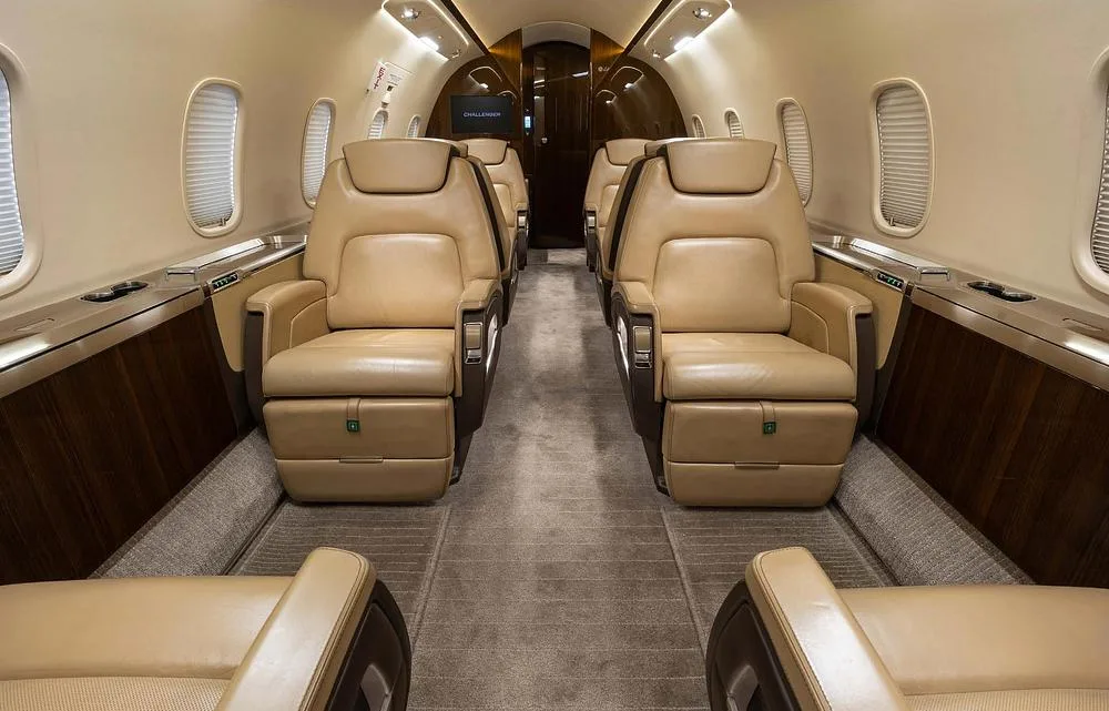 A view down the aisle of a Challenger business jet cabin, showing multiple rows of beige leather seats and dark wood accents.