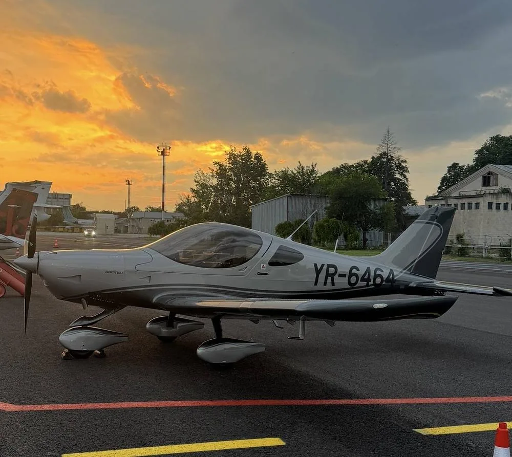 A silver and dark grey Pipistrel Virus SW aircraft (YR-6464) parked on an airfield with a vibrant sunset sky.