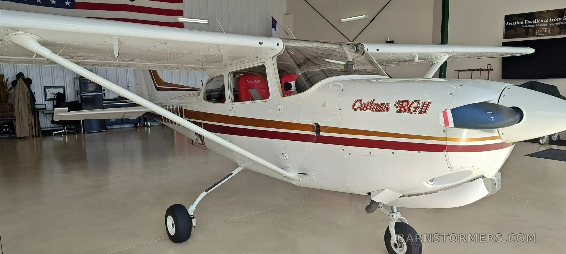 A white Cessna Cutlass RGII (N5393V) with red and gold stripes is shown from a front-left angle inside a hangar.