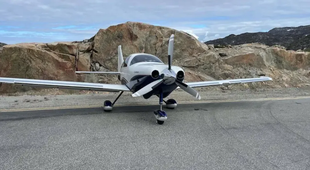 A white and blue Columbia 400 aircraft with a three-bladed propeller parked on an asphalt ramp with rocky terrain in the background.