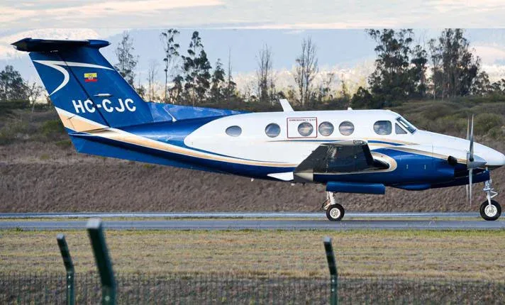 A blue and white Beechcraft King Air HC-CJC on a runway with trees in the background.