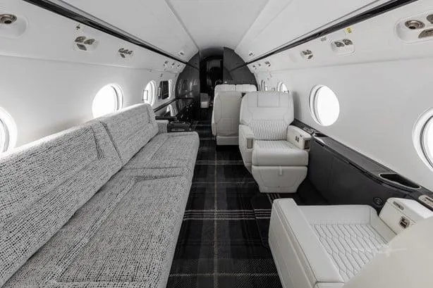 The interior of a private jet cabin, showing a long grey sofa and individual white seats with a dark plaid carpet.