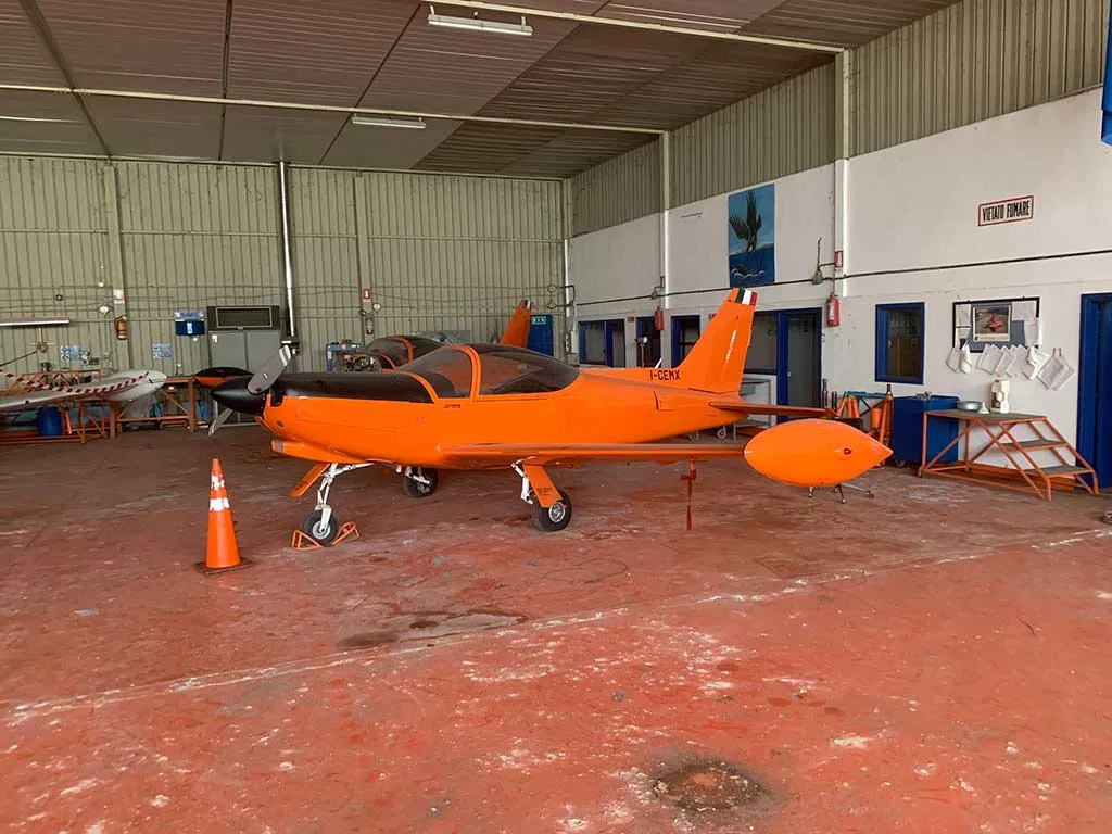 An orange and black single-engine aircraft, possibly a Falco F.8L, is parked inside a hangar with a concrete floor.