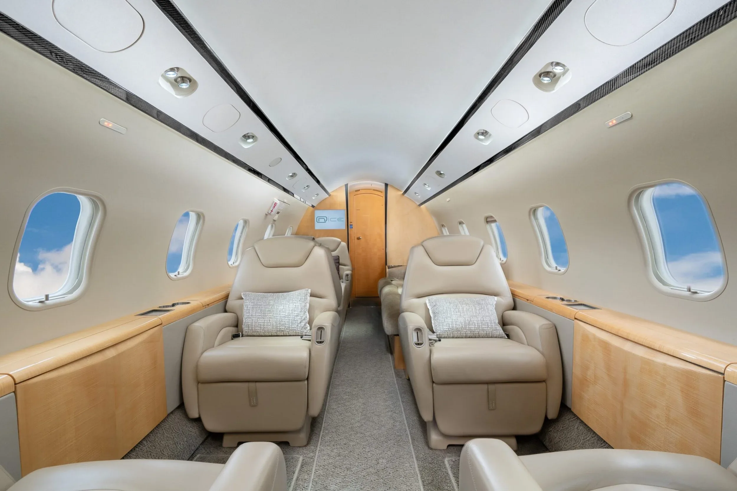 The interior of a private jet cabin with light beige leather seats and light wood paneling, looking towards the rear, with pillows on the seats.