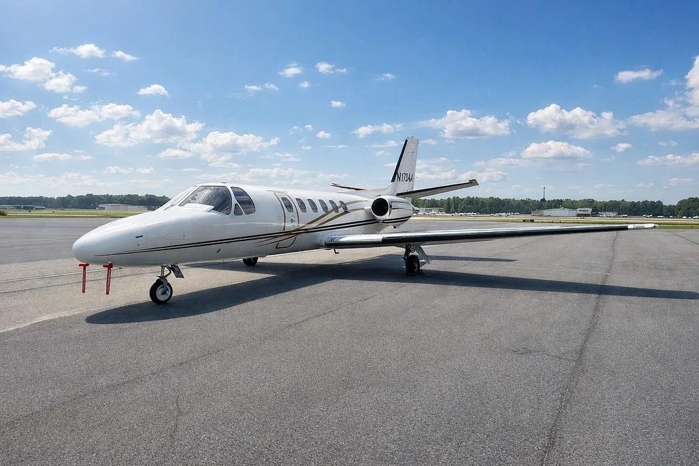 A white business jet with gold and black stripes, tail number N173AA, viewed from the front-left on an airfield under a blue sky with clouds.