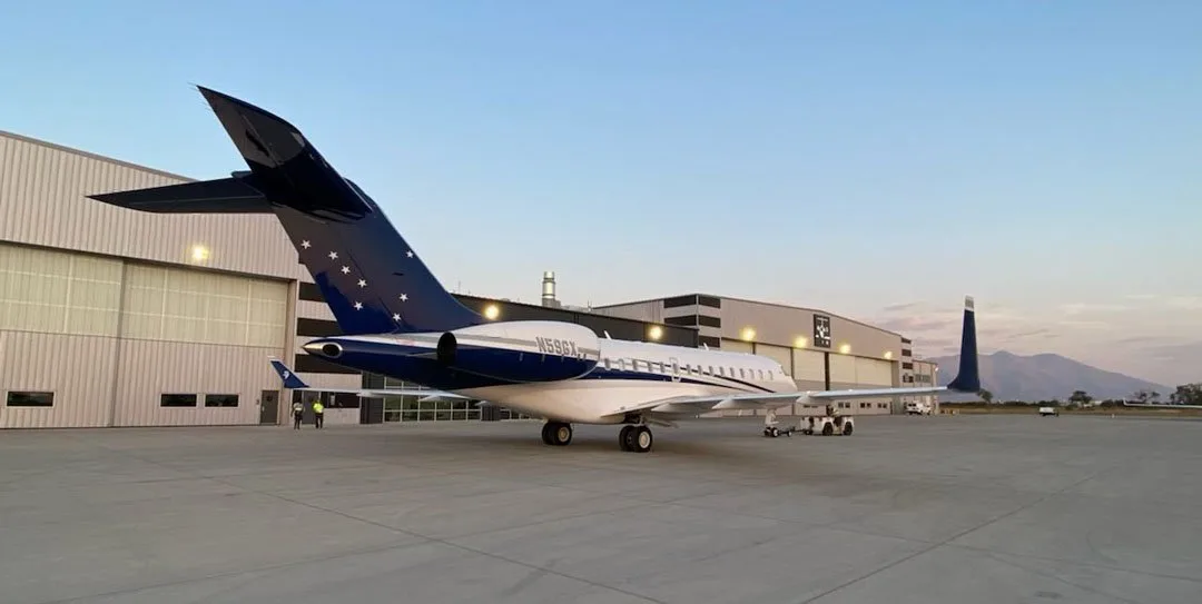 A white and blue Bombardier Challenger 605, N59GX, is parked on a tarmac next to hangars at dusk.