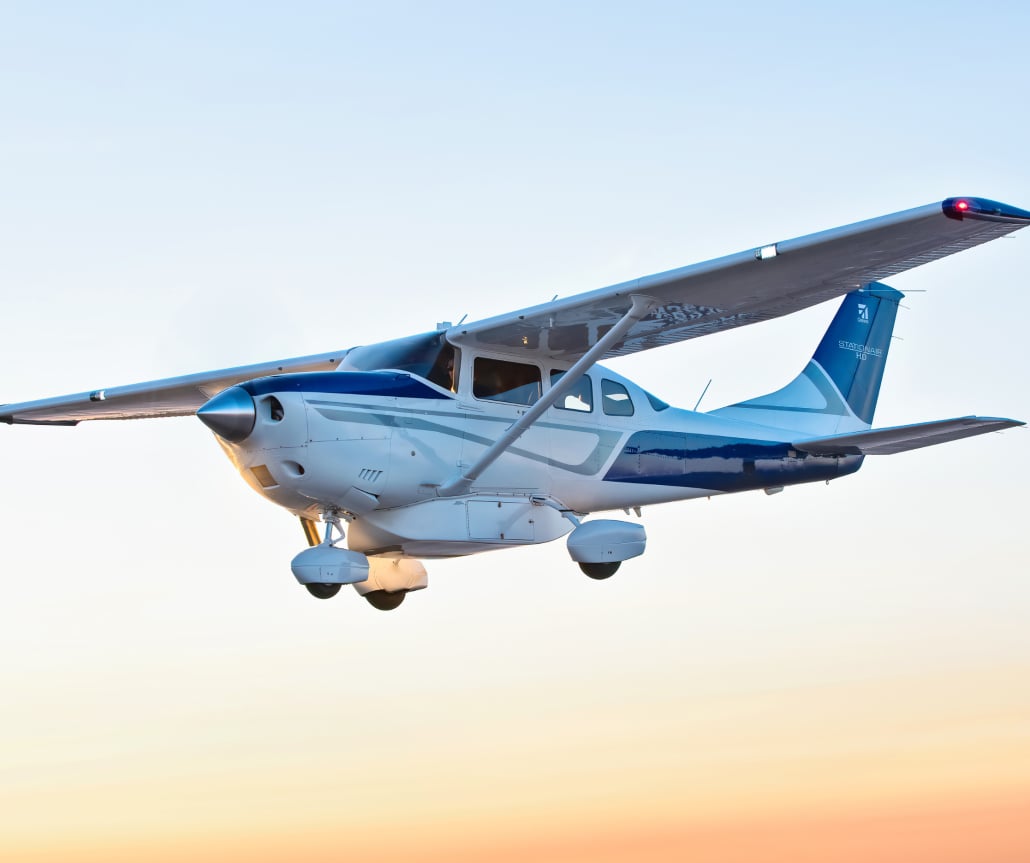 A high-wing single-engine aircraft is in flight against a colorful sky.
