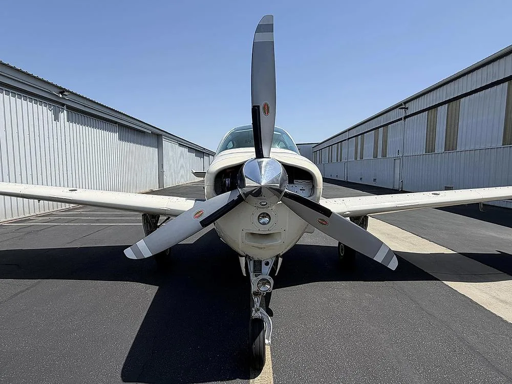 A head-on view of a white and gold Beechcraft Bonanza, highlighting its propeller and nose landing gear.