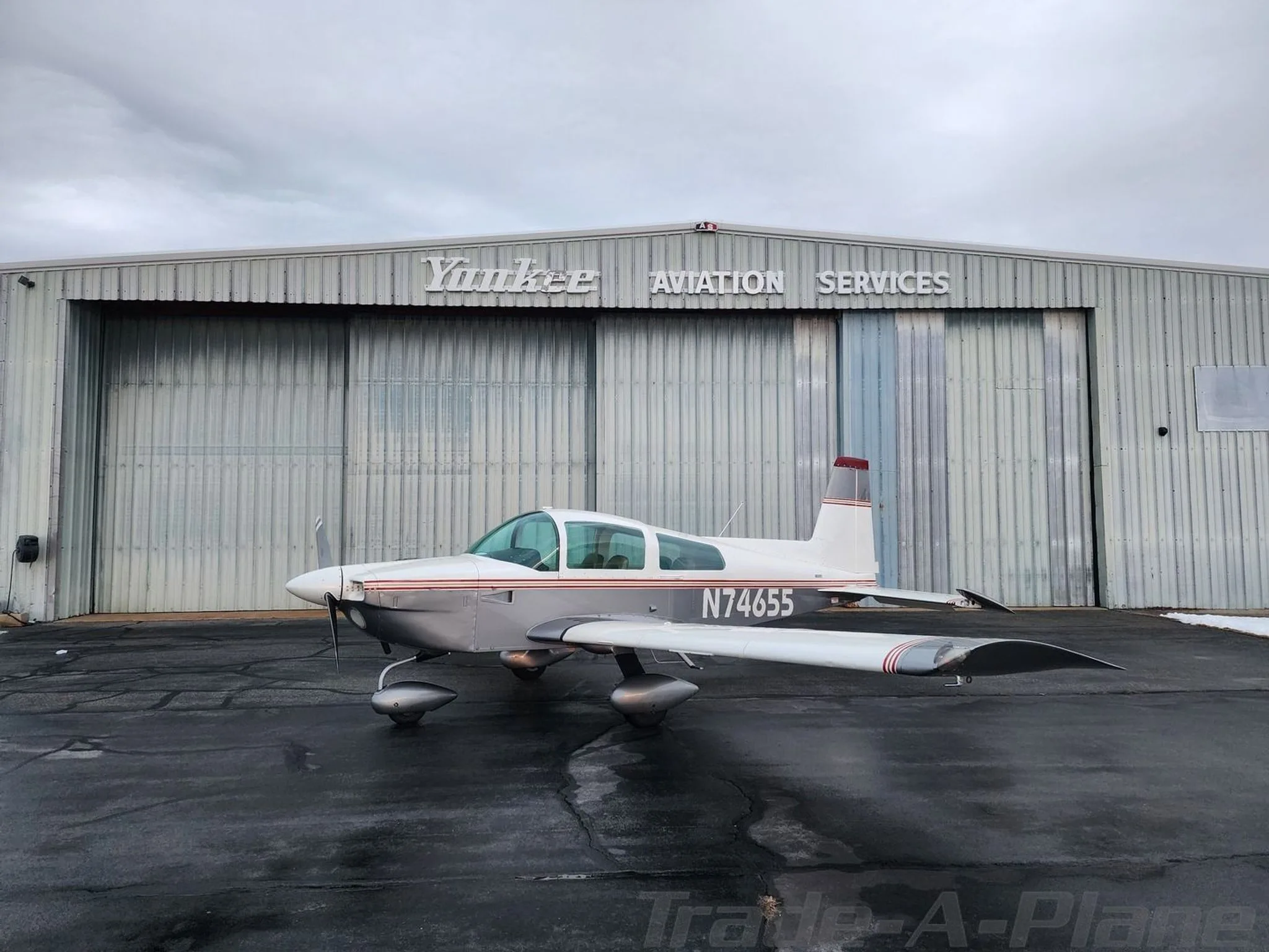A white and grey Grumman American aircraft, N74655, parked on a wet tarmac in front of a "Yankee Aviation Services" hangar under a cloudy sky.