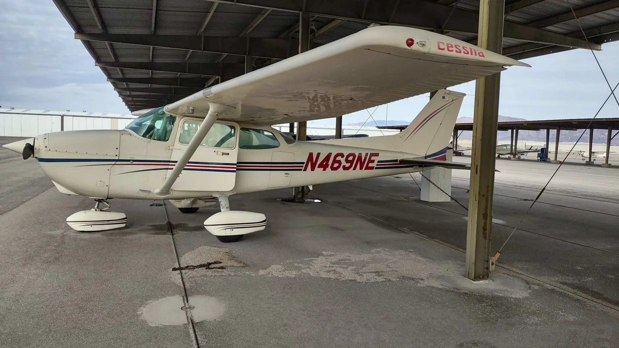 A white Cessna 172 with blue and red stripes parked under a hangar, showing the left side and tail.