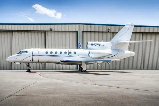 A white Dassault Falcon 900EX business jet with blue stripes, registration N47NS, parked in front of a hangar.