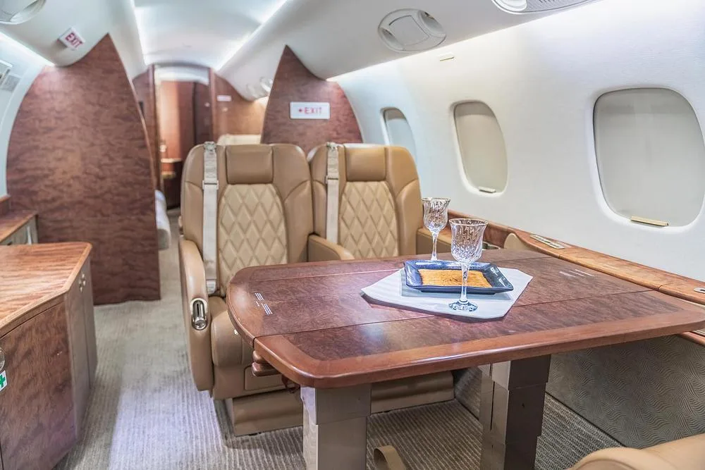 A dining area within the business jet cabin features a polished wood table set with two crystal glasses and tan leather seats.