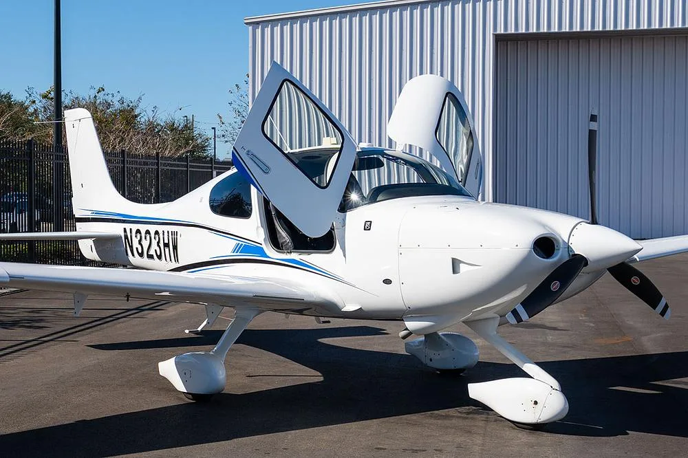 A white Cirrus SR22 aircraft with blue and black stripes, registration N323HW, parked with its gull-wing doors open next to a metal hangar.