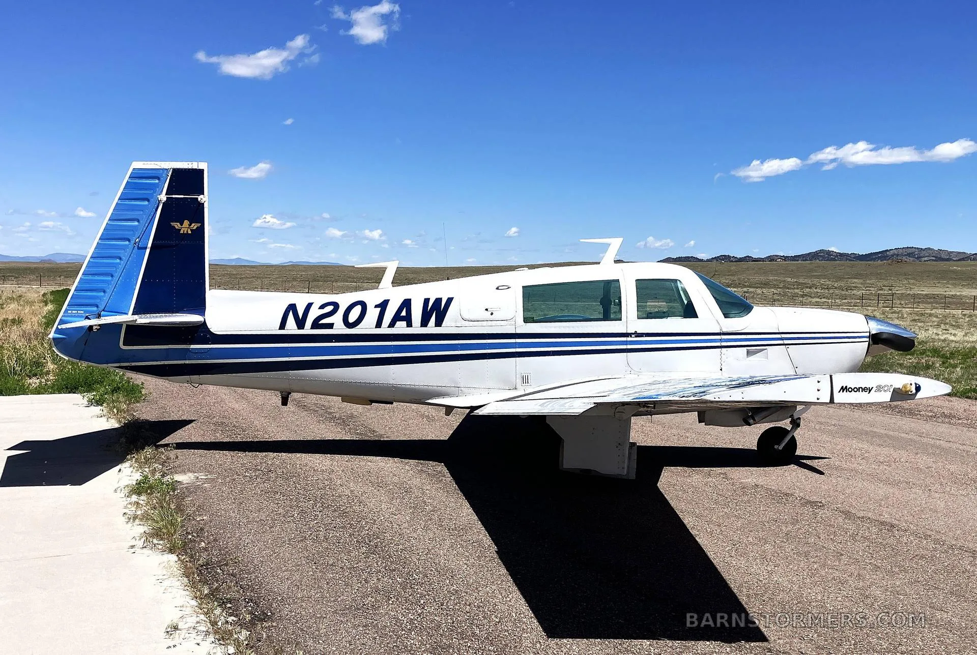 A white Mooney M20J aircraft with blue stripes and tail, registration N201AW, parked on an asphalt runway under a clear blue sky.