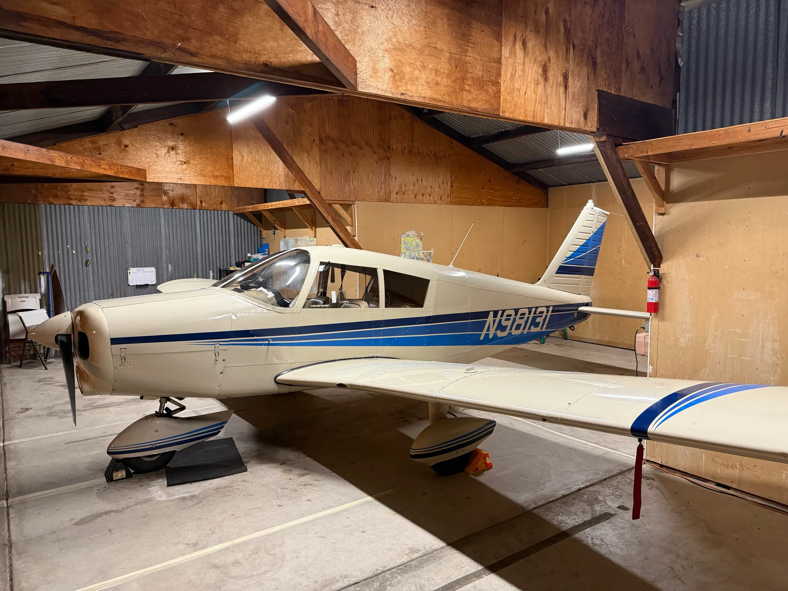 A white and blue Piper Cherokee aircraft (N98131) is parked inside a wooden hangar.