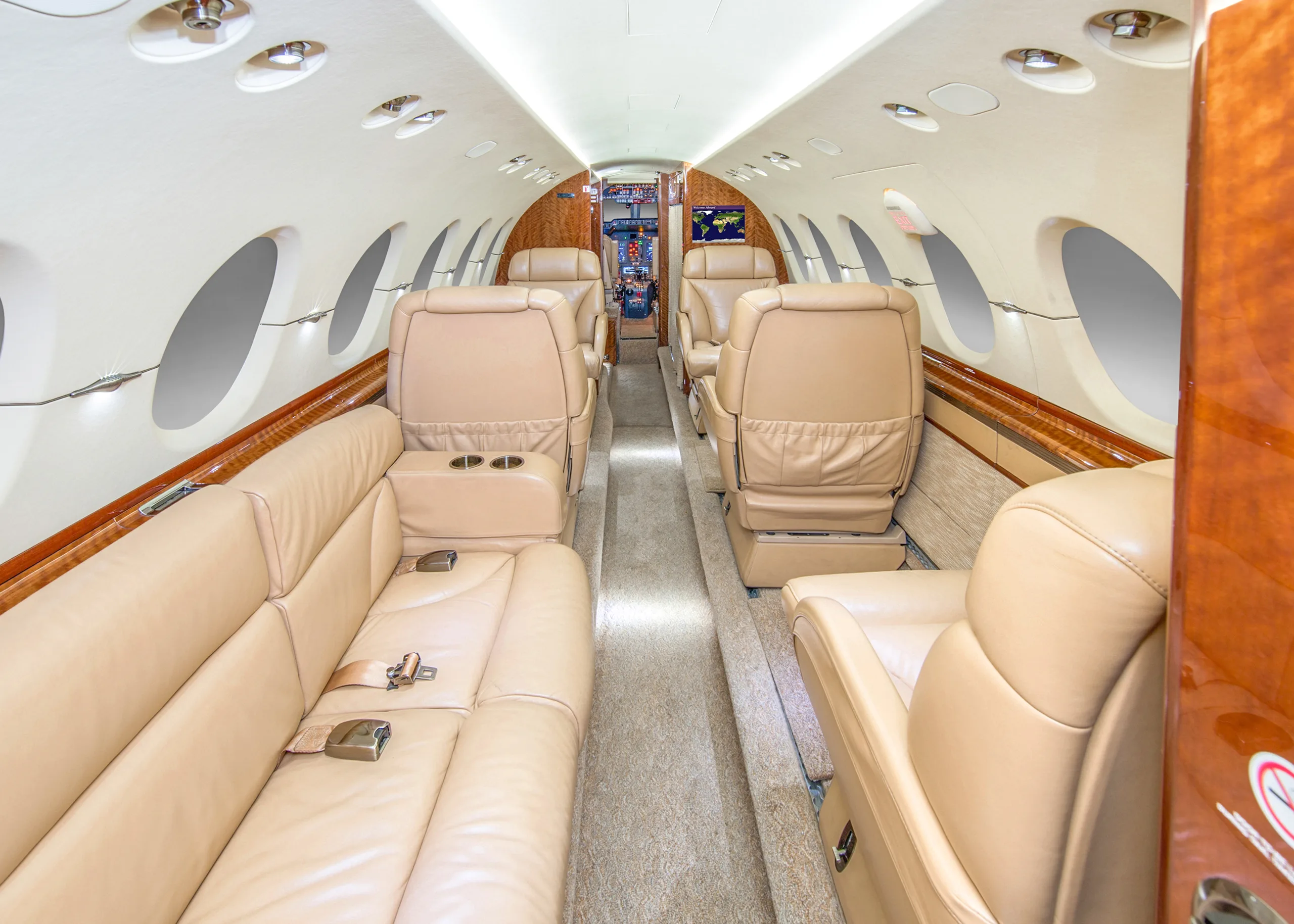 The spacious interior of a private jet cabin featuring a long tan leather sofa, individual seats, and wooden accents.