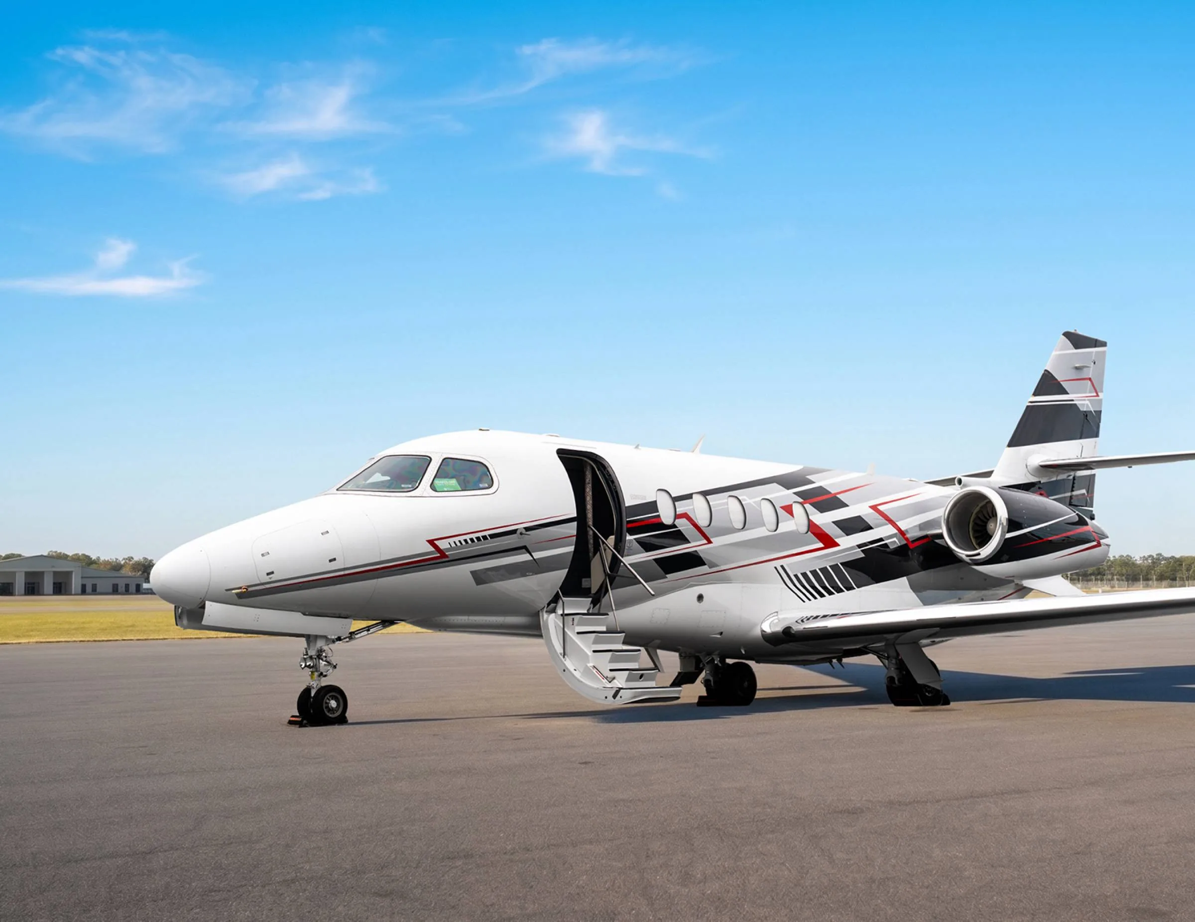 A white business jet with black, red, and grey geometric livery is parked on an asphalt runway, viewed from the front-left, with its door open and stairs extended.