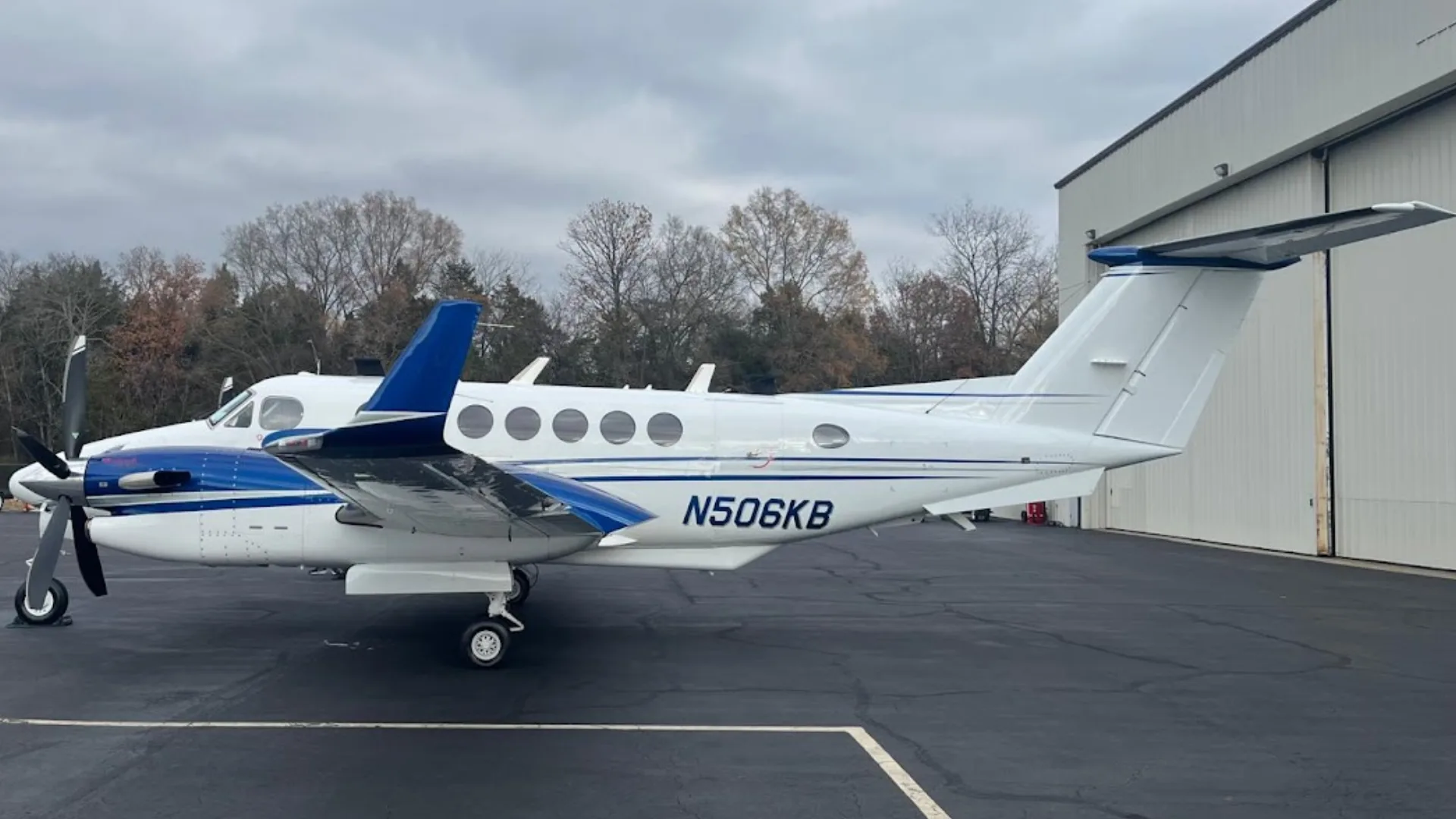 A white and blue Beechcraft King Air N506KB is parked on an asphalt ramp next to a hangar under a cloudy sky.