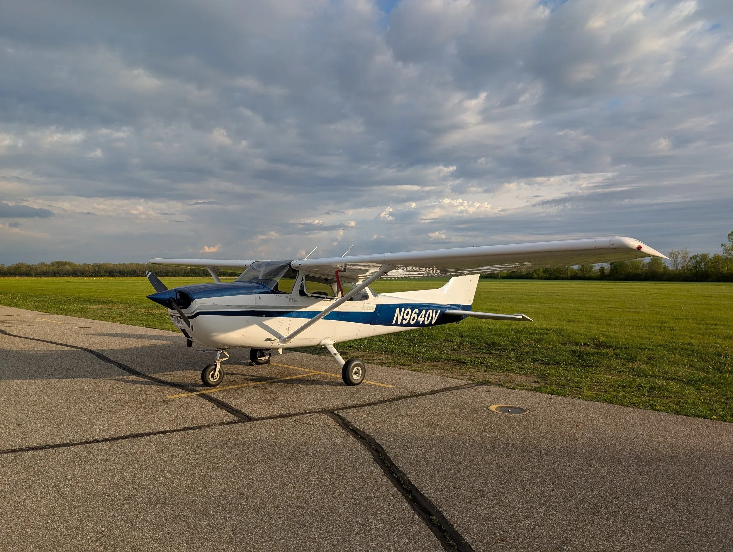 A white and blue Cessna 172 (N9640V) is parked on an asphalt ramp next to a green field under a cloudy sky.