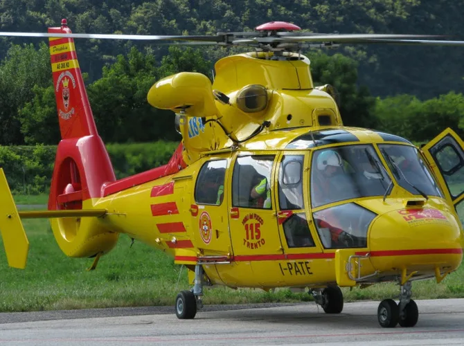 A yellow and red Airbus Helicopters Dauphin AS365 N2 emergency services helicopter with '115 TRENTO' markings on an airfield with trees in the background.