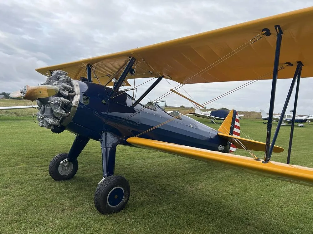 A blue and yellow biplane with a radial engine is parked on a grassy field under a cloudy sky, showing the N60955 registration on the tail.