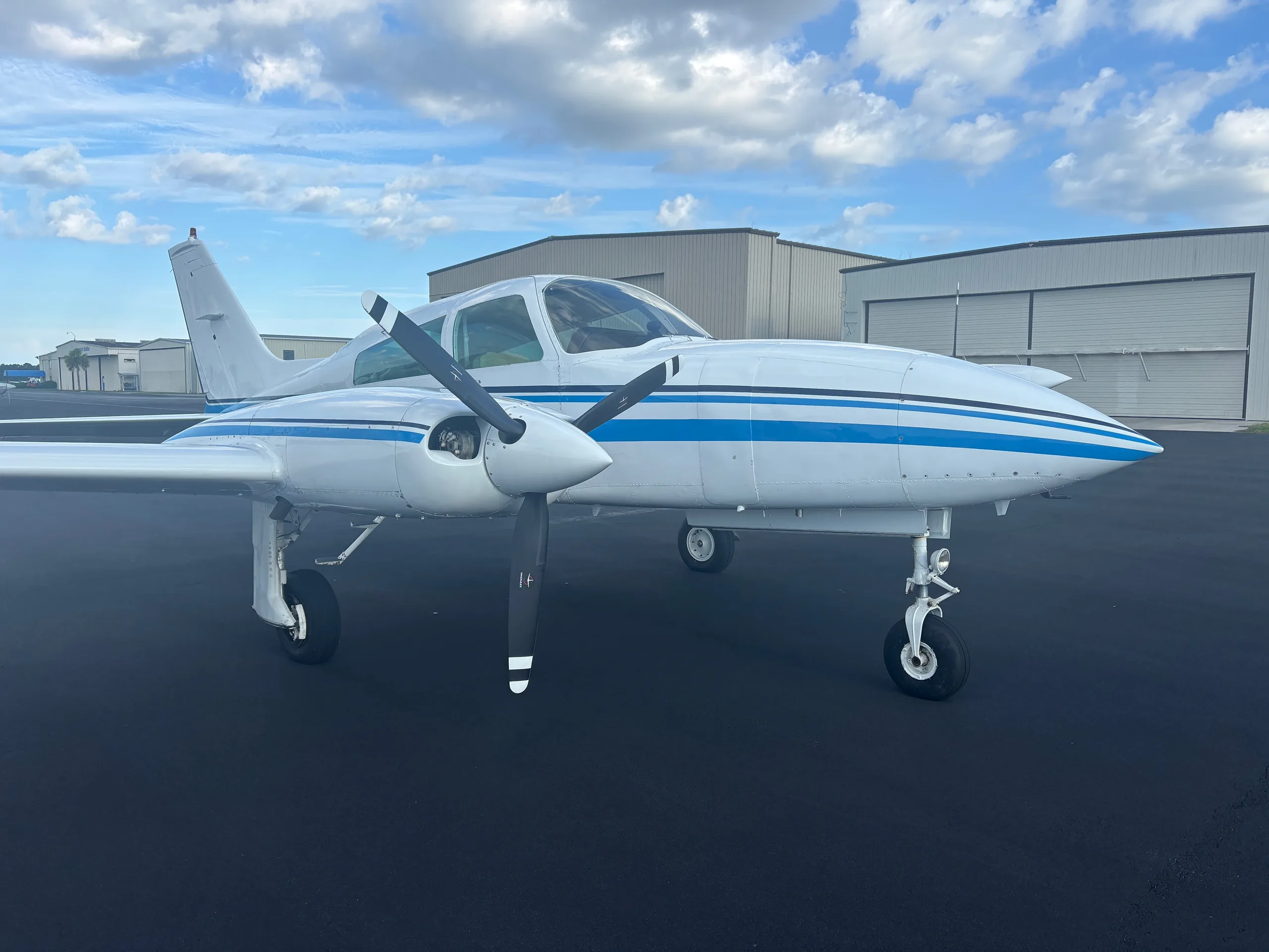 A white twin-engine aircraft with blue stripes on the tarmac, viewed from the front-left with hangars in the background.