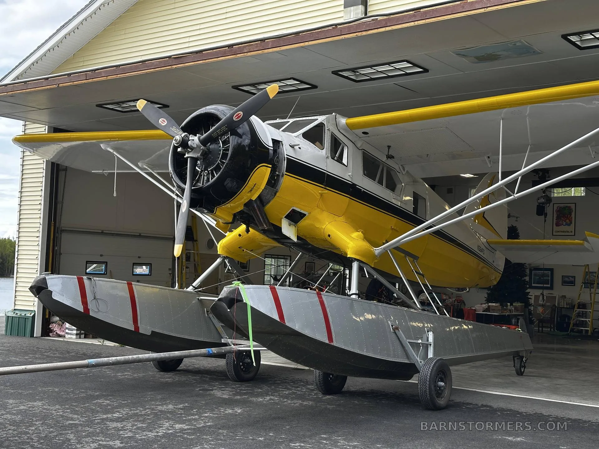 A yellow and white floatplane on a dolly is being pulled out of a hangar, showing its large floats and propeller.