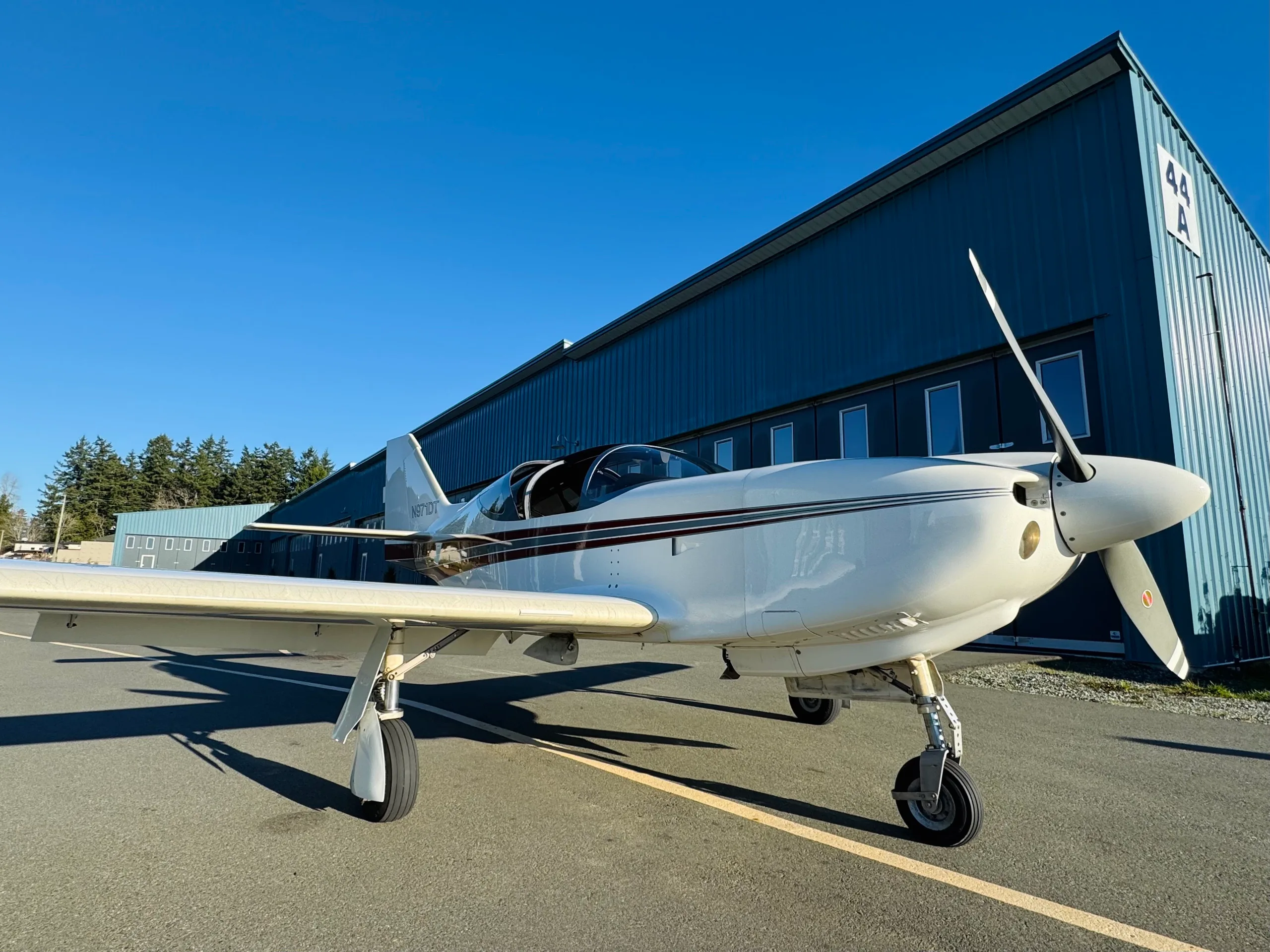 A white single-engine aircraft with brown and black stripes, registration N971DT, parked on an tarmac in front of a blue hangar on a sunny day.