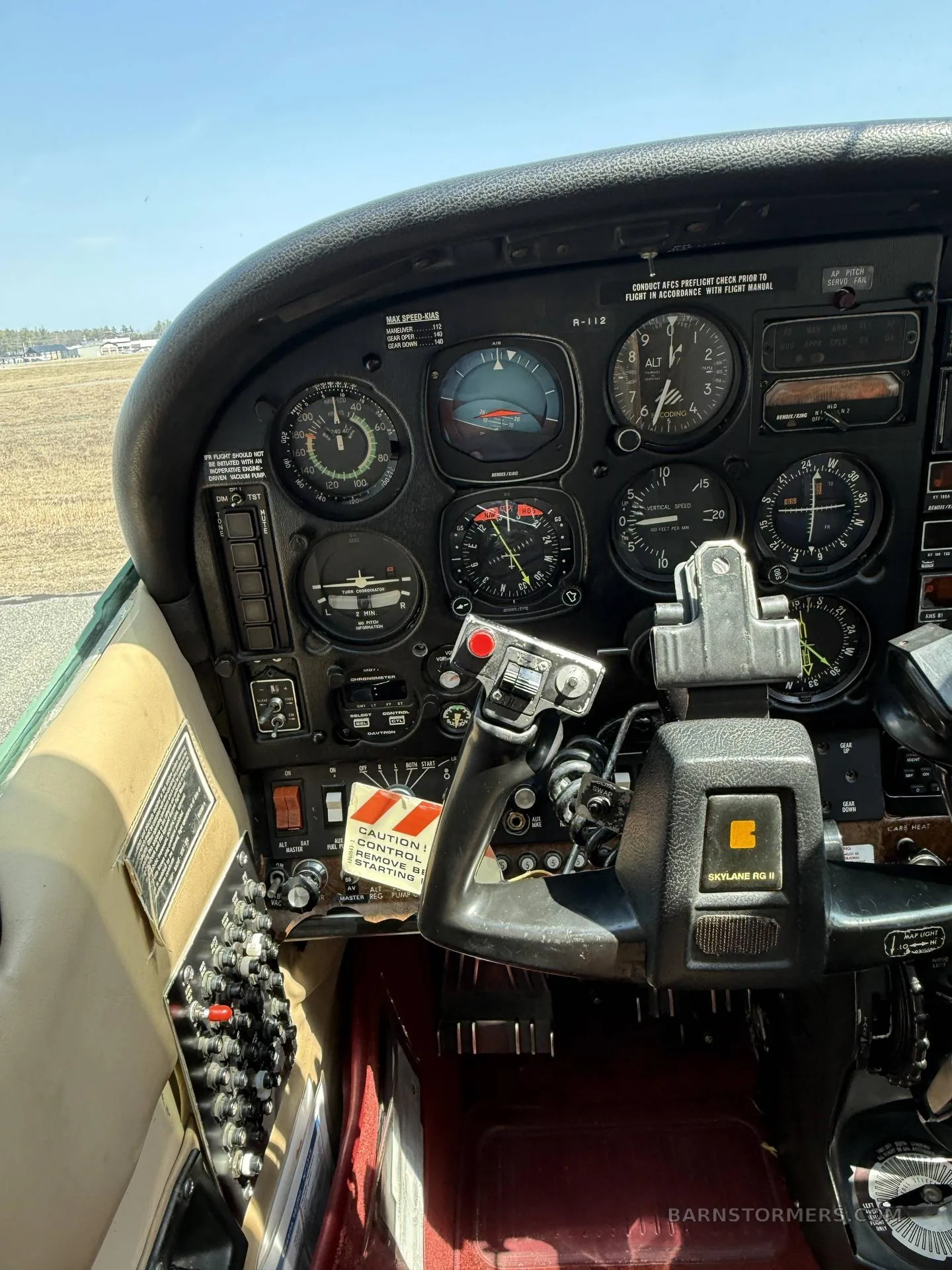 The pilot's side of a light aircraft cockpit, featuring the instrument panel, yoke, and a view of the airfield outside.