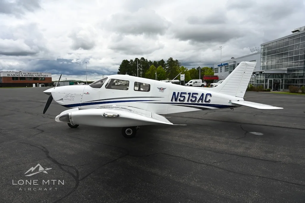 A white single-engine propeller aircraft with blue accents, registration N515AC, parked on an asphalt tarmac next to a modern building.