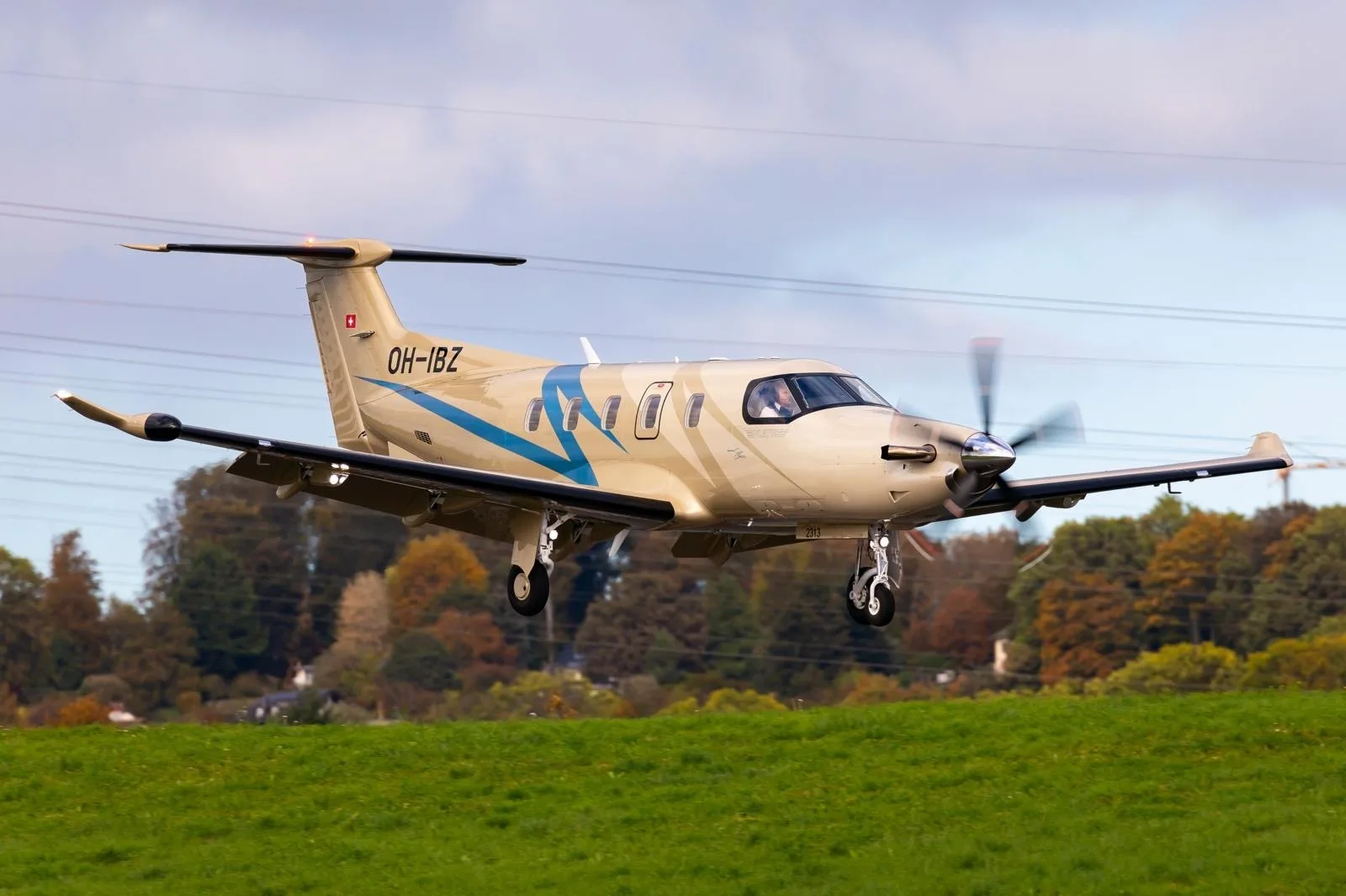 A tan and blue Pilatus PC-12 turboprop aircraft with registration OH-IBZ is landing over a green field.