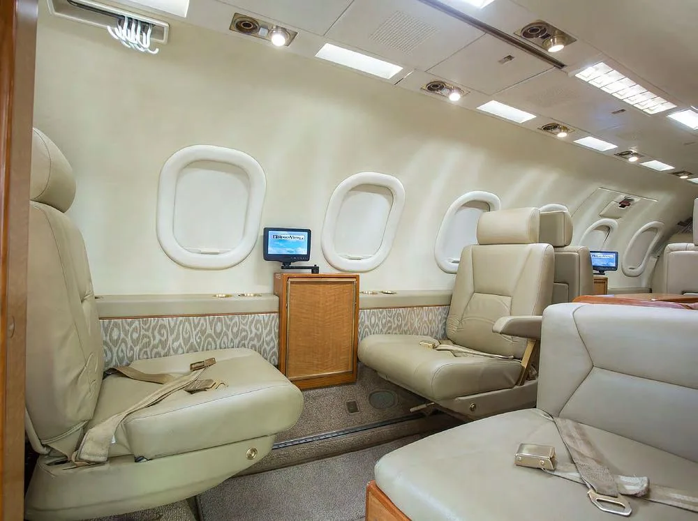 The aircraft cabin interior shows multiple beige leather seats, windows, and a small entertainment monitor.