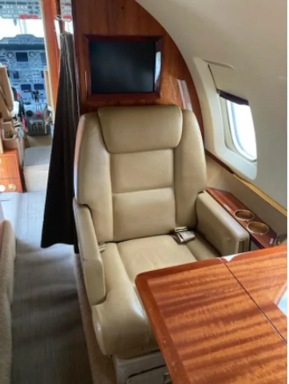 A tan leather cabin seat with a wooden table and monitor, looking towards the cockpit of an aircraft.
