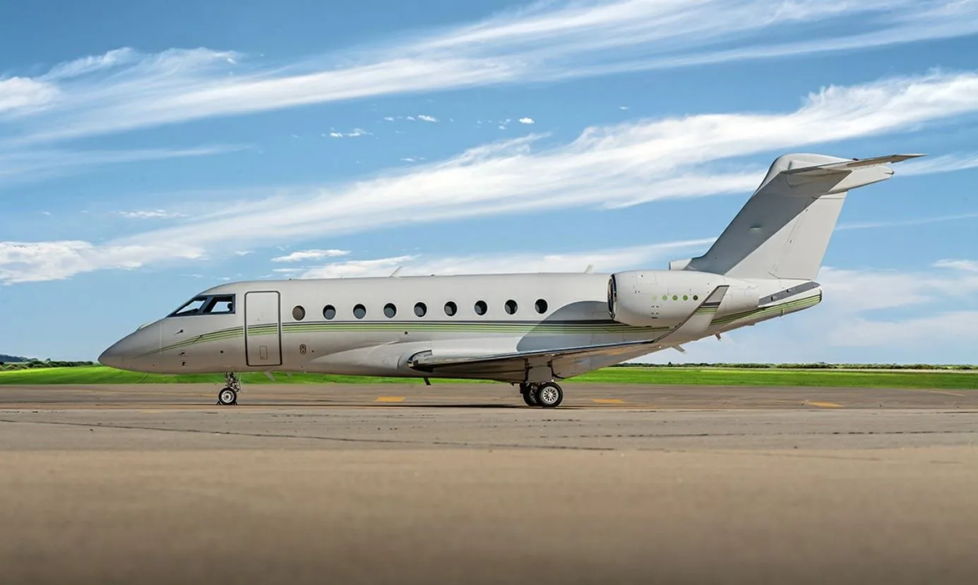 A light grey business jet with a green stripe on the fuselage, parked on an airfield under a blue sky.