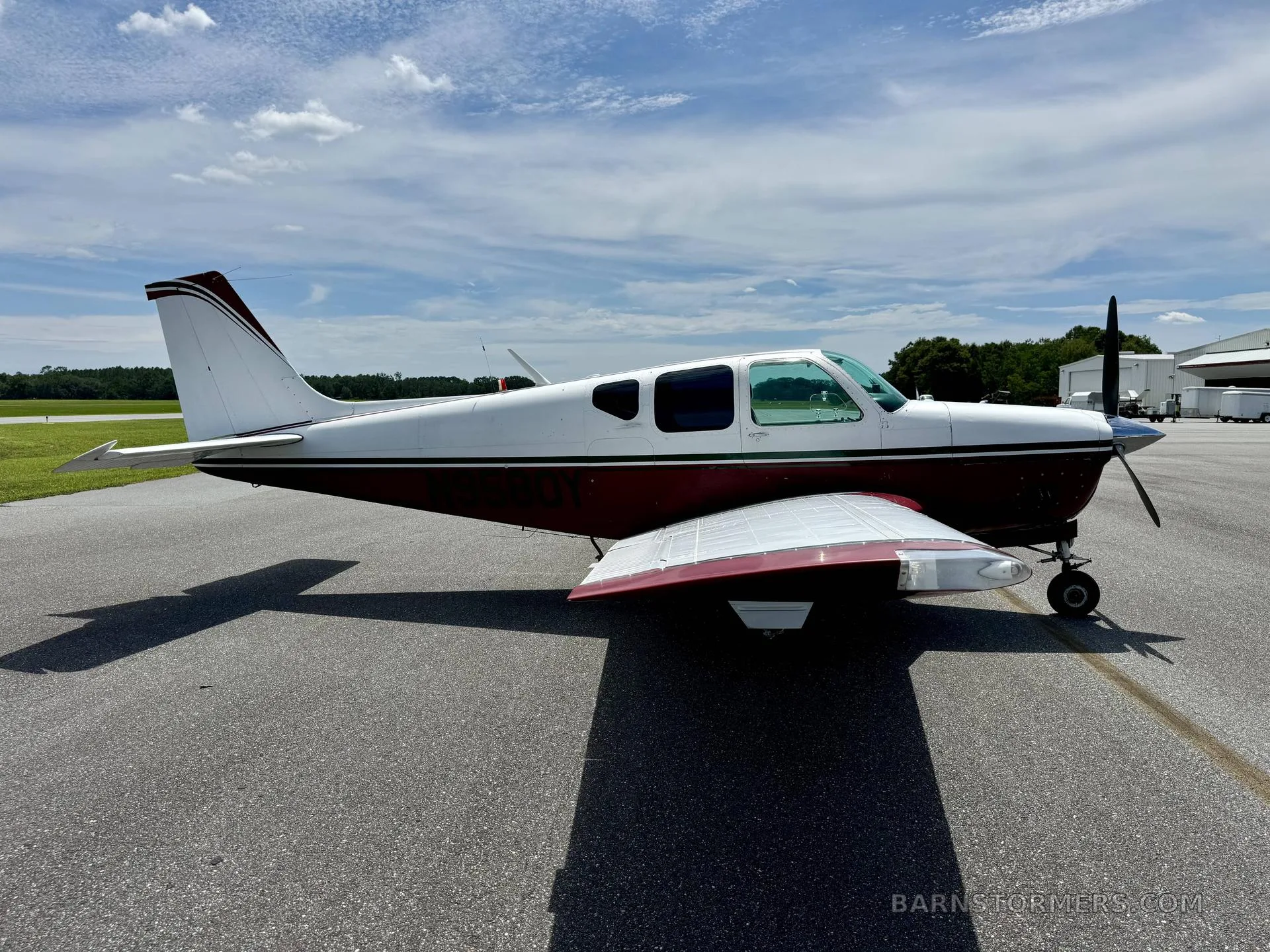 A white and maroon single-engine propeller aircraft is viewed from the side on an airfield under a sunny sky.