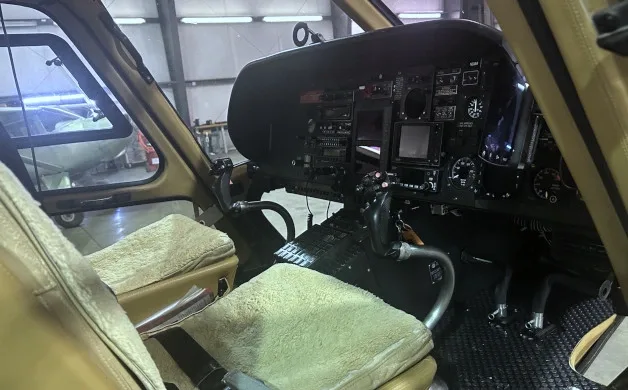 The cockpit of a helicopter with tan sheepskin seat covers, showing the instrument panel and controls.