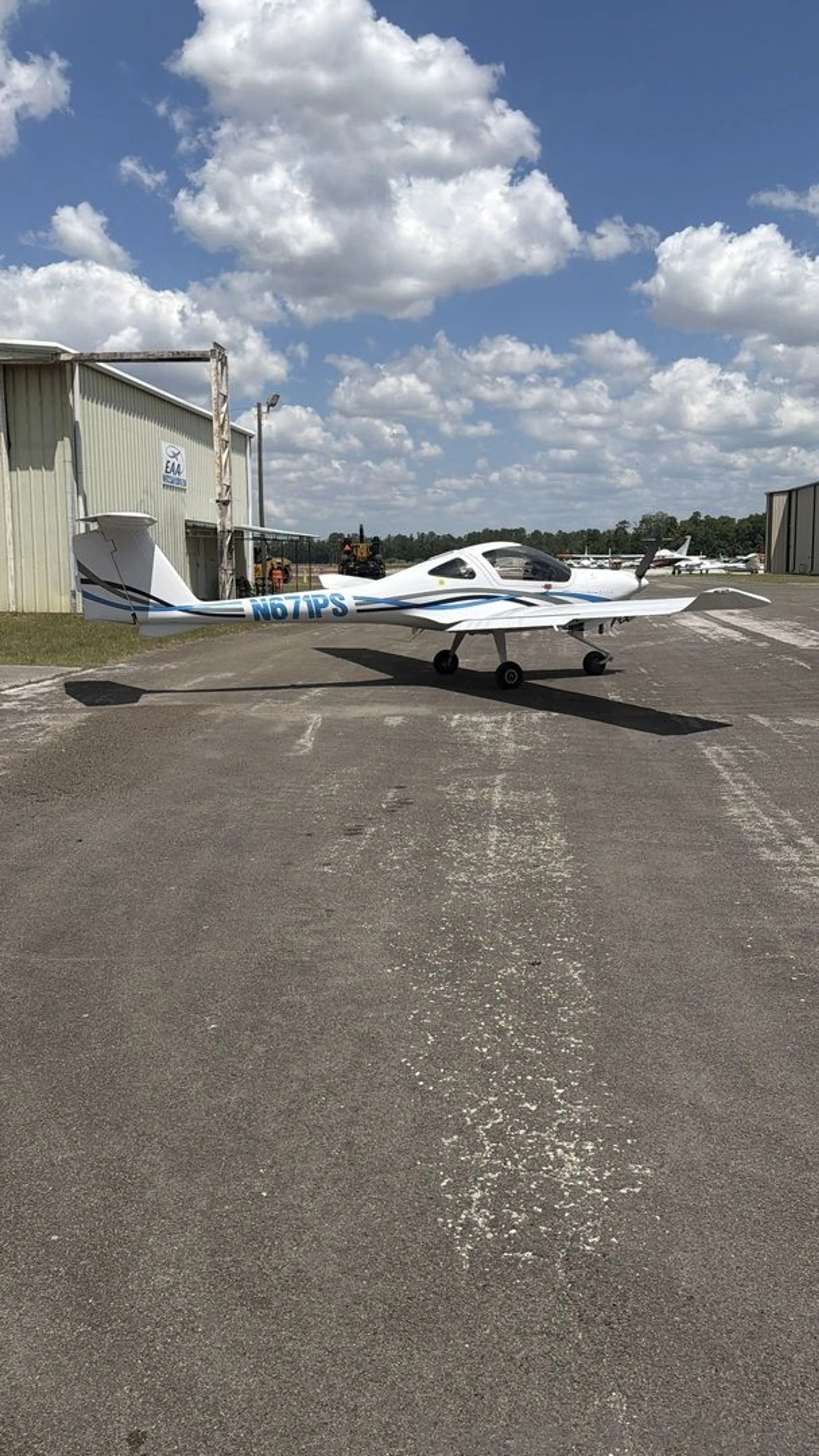A white Diamond DA20-C1 Eclipse aircraft with blue stripes, registration N671PS, parked on an asphalt tarmac with hangars in the background under a cloudy sky.