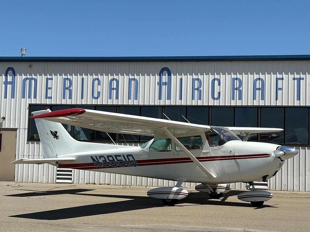 A white Cessna aircraft with red stripes and N3951Q registration parked in front of the American Aircraft building, viewed from the front-right.