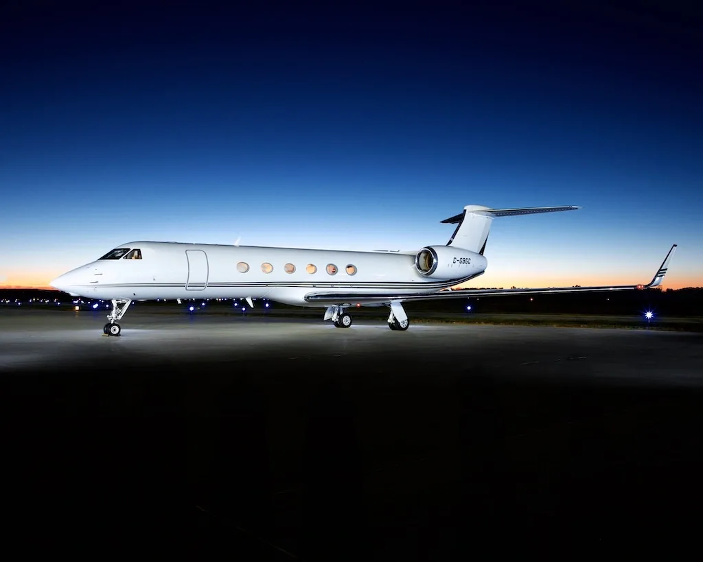 A white Gulfstream G650 business jet with registration F-GBSC parked on a tarmac at dusk, illuminated by ground lights.